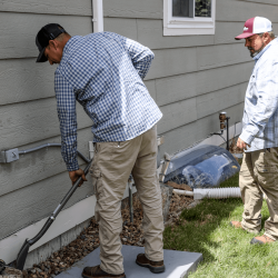 The image shows two HVAC experts outside creating a clearing for a new HVAC unit.